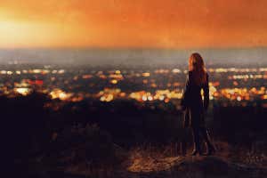 Vista trasera de una mujer con pelo largo rojo vistiendo abrigo negro y botas de tacón de pie en la colina viendo la puesta de sol sobre el valle de las luces de la ciudad